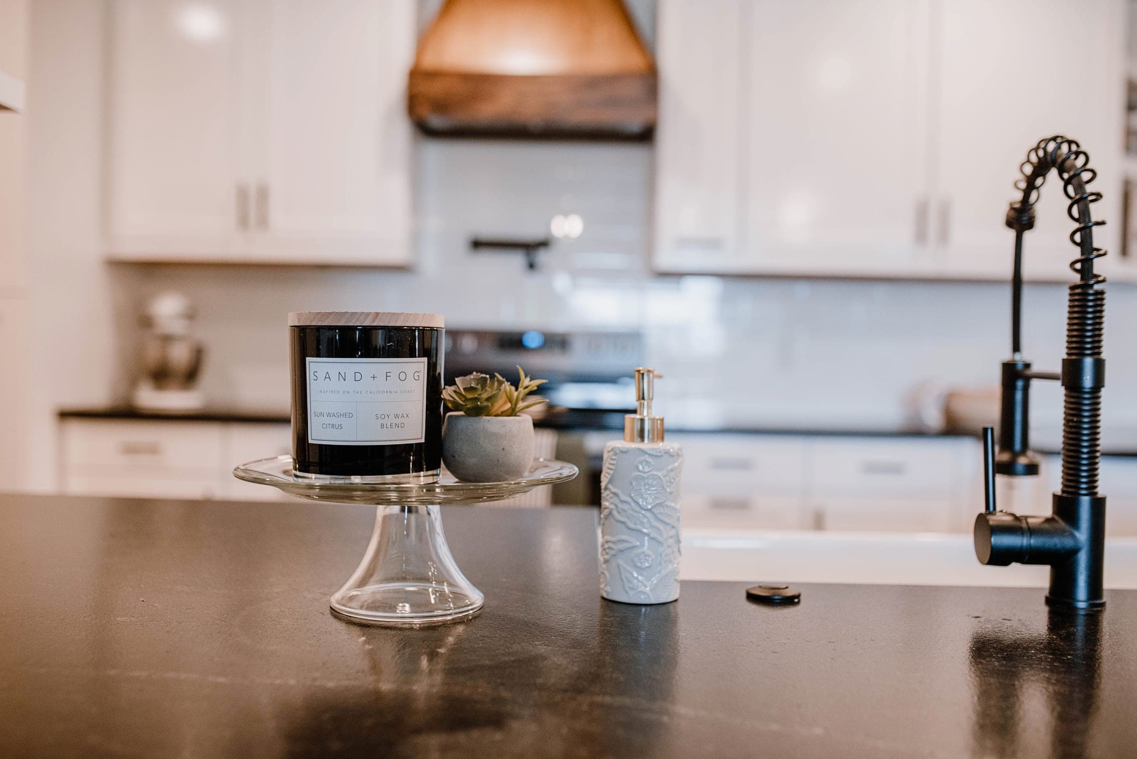 Finished kitchen detail with a candle and succulent on a dark countertop, white cabinetry and a wood range hood behind
