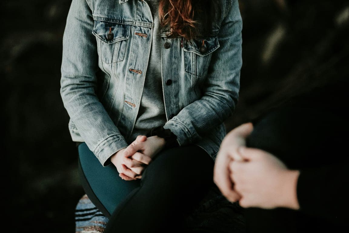 Person seated with hands clasped in a quiet counseling session, another persons hands in the foreground