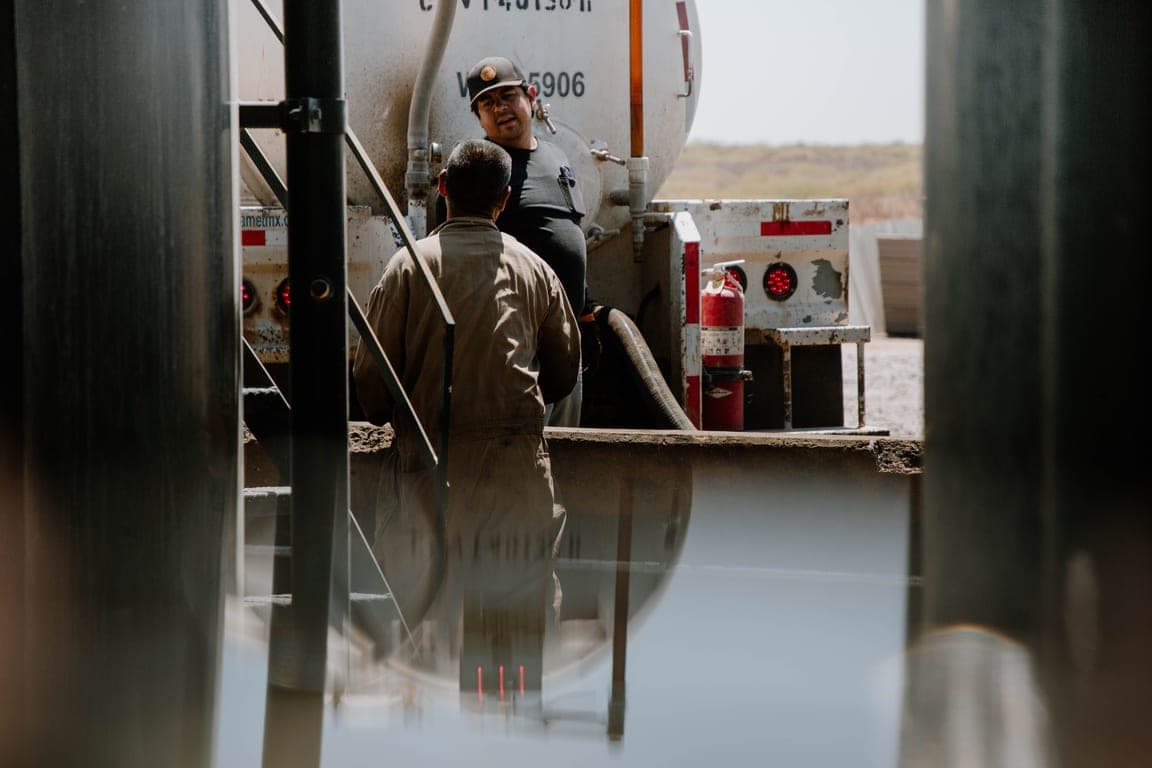 Two oilfield workers in coveralls talking beside a tanker trailer at a processing site