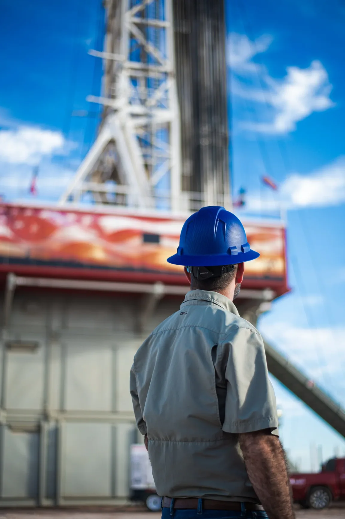 Worker in a blue hard hat looking up at a tall drilling rig derrick against a bright blue sky