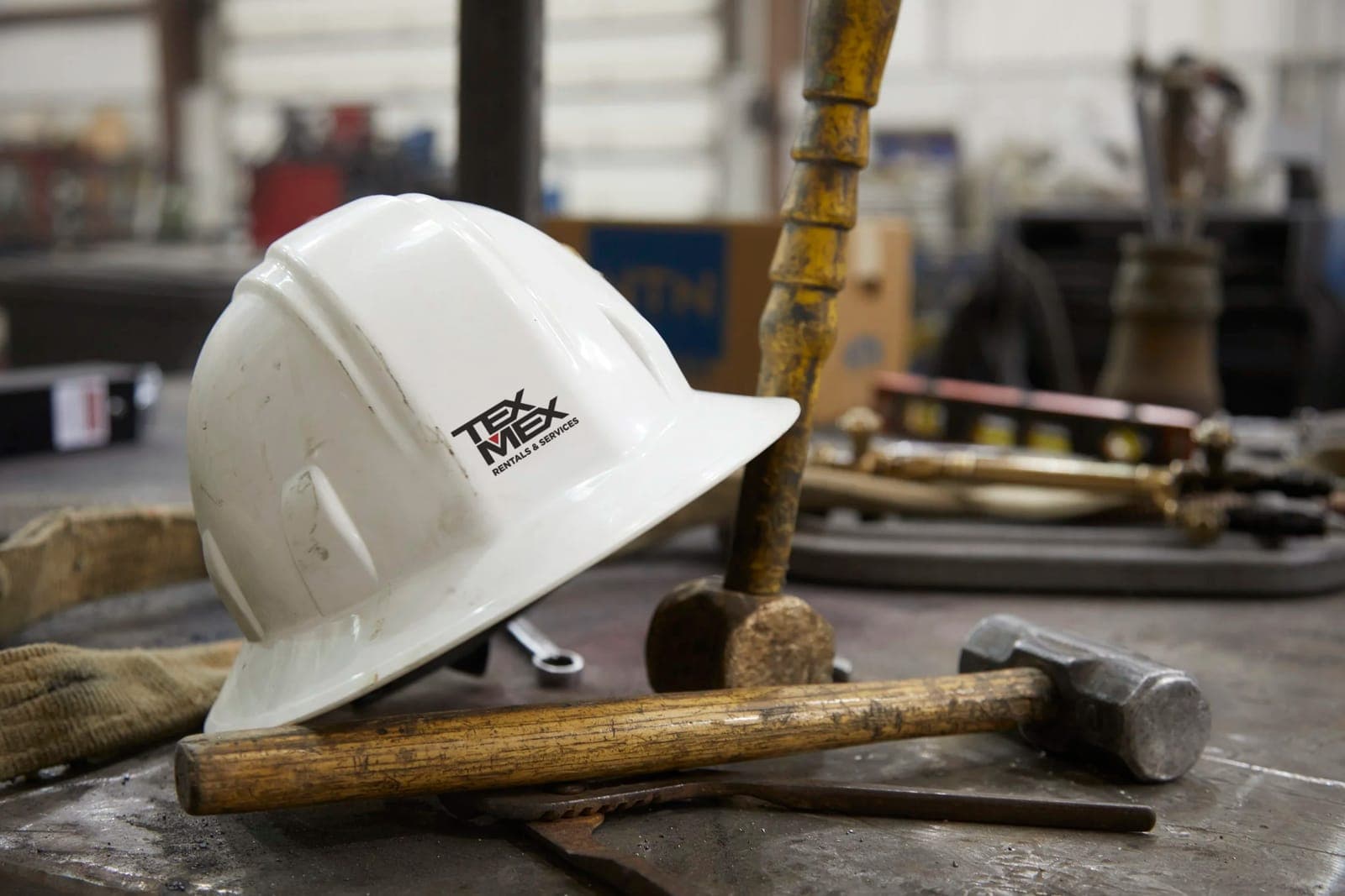 White hard hat with the Tex Mex Rentals & Services logo resting on a steel workbench beside sledgehammers and wrenches